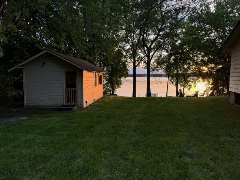 Sunset over a lake with a yellow cabin in the foreground and wooden chairs by the water.