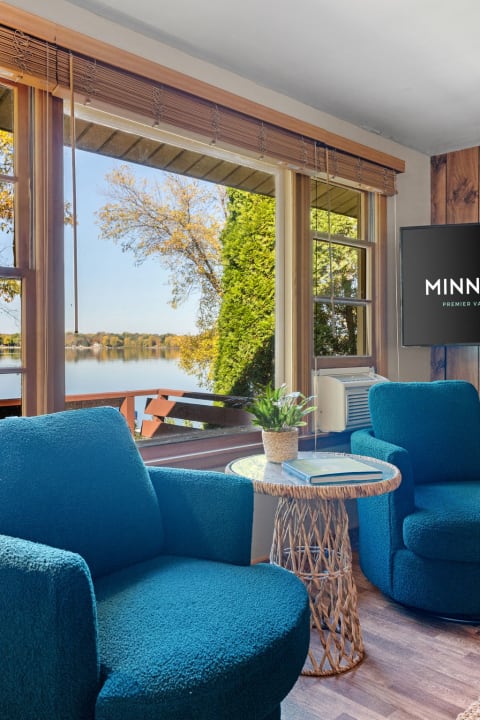 This image depicts a living space with turquoise chairs, a round rattan coffee table, and large windows overlooking a tranquil lake.