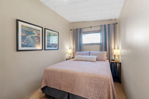 A cozy bedroom featuring a pink bedspread, framed artworks, and lamps.