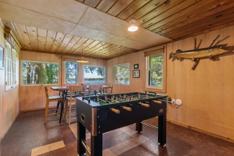 Interior of a wooden cabin with a foosball table and large windows showing nature outside.