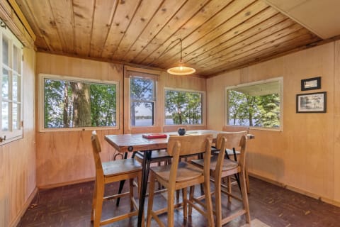 Interior of a lakeside room featuring a wooden table, chairs, and views of trees and water through large windows.