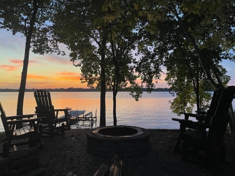 Silhouette of Adirondack chairs near a lake at sunset with a fire pit.