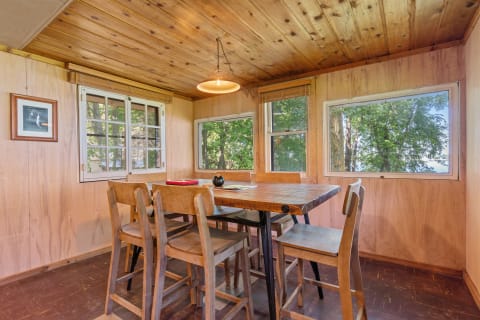 Interior view of a rustic cabin's dining area with wooden furniture and large windows overlooking trees.