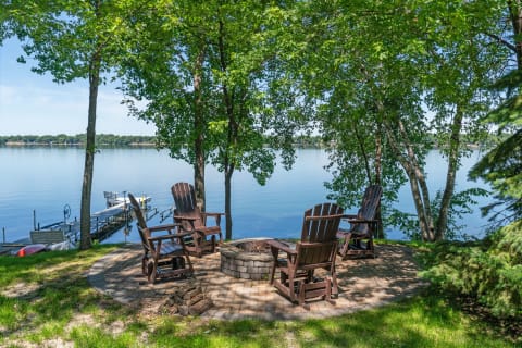 Lakeside patio with Adirondack chairs surrounding a fire pit, overlooking calm water and a dock.