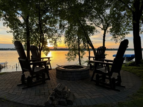 Wooden rocking chairs by a lake at sunset, surrounded by trees.