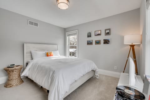 A modern bedroom with a queen-sized bed, decorative pillow, woven accent table, and framed photos on the wall.