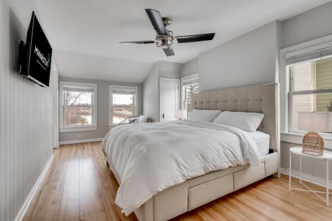 A cozy modern bedroom featuring a beige tufted bed, light wood floors, and ample natural light from windows.