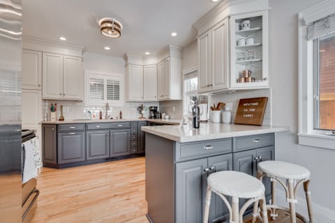 A cozy kitchen with dark gray and white cabinets, a welcoming sign, and bar stools.