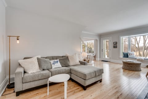 A bright living room featuring a gray sofa, wooden floors, and large windows overlooking trees.