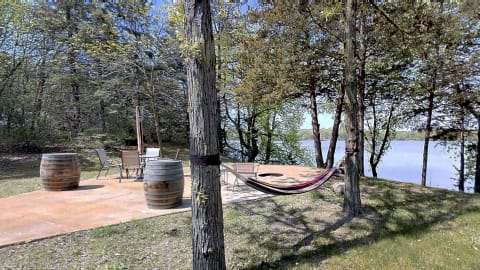 Lakeside patio featuring a colorful hammock, chairs, and wooden barrels under a tree canopy.