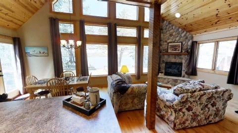 Interior of a cabin featuring large windows, a floral couch, and a dining area with a scenic lake view.