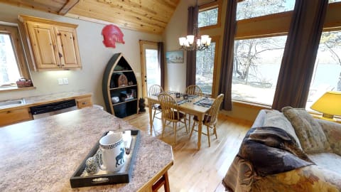 Interior view of a rustic cabin with a kitchen and dining area, featuring large windows overlooking nature.
