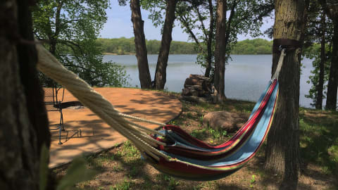 A hammock hangs between two trees beside a calm lake, with greenery and a wooden platform in view.