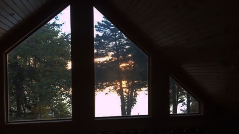 A cozy cabin window with a view of tall trees and a sunset in the background.