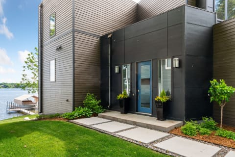 A modern entrance to a lakeside home featuring a black wall, blue door, and lush landscaping.