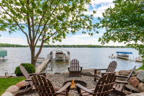 View of a lakeside with Adirondack chairs around a fire pit, a dock with a boat, and a canopy of trees.