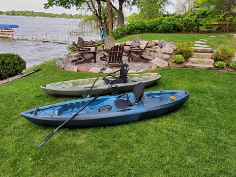 Two kayaks resting on the grass near a tranquil lake with Adirondack chairs in the background.
