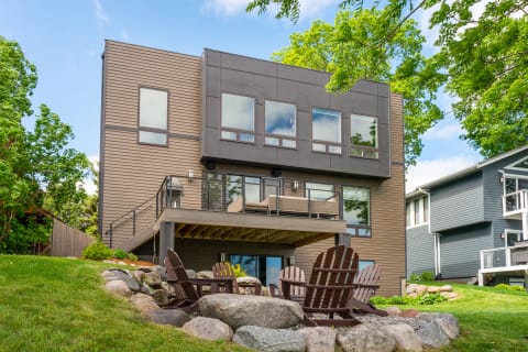 A modern two-story house with a balcony, Adirondack chairs, and a landscaped yard.