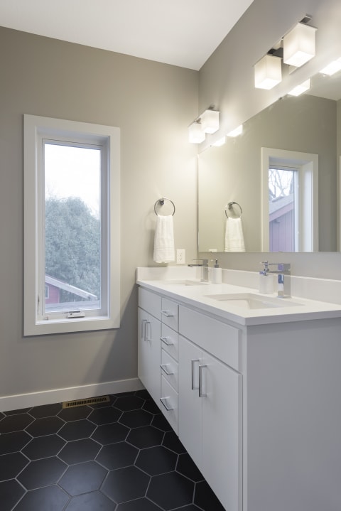 A contemporary bathroom with a white double sink vanity, gray walls, and a black hexagonal tiled floor