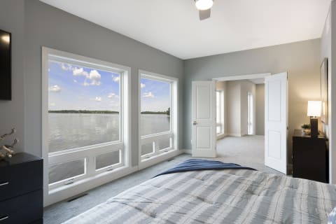 A serene bedroom with large windows overlooking a lake, featuring a bed and dark dresser.
