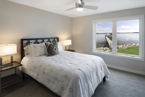 A cozy bedroom with a bed, nightstands, and a large window showing a lake with a dock.