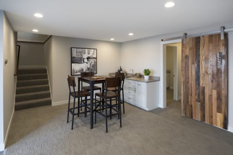 A sleek basement space featuring a high-top dining table, kitchenette, and a wooden barn door.