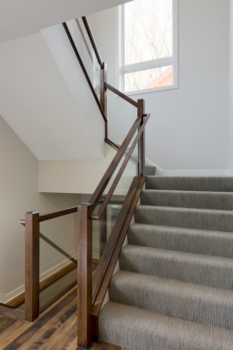 Interior view of a staircase featuring gray carpet and a sleek wooden handrail with glass panels.