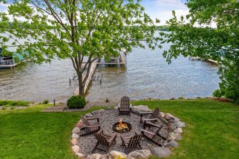A circular fire pit with Adirondack chairs, near a lake and dock.