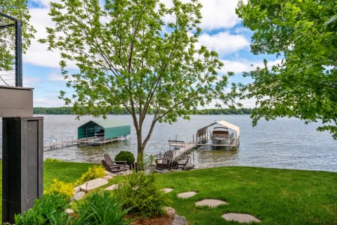Lakeside scene featuring a dock with boats and Adirondack chairs on green grass.