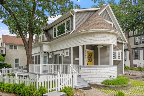 Exterior view of a classic house featuring gray siding, white shingles, a round porch, and a yellow front door.