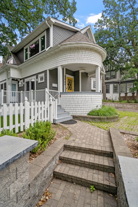 A house featuring gray siding, a white shingle porch, and a bright yellow door surrounded by greenery and a white fence.