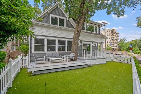 A cozy gray house with a porch, surrounded by a green lawn and fence, set against a blue sky.