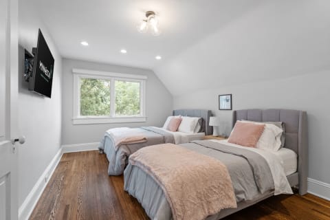 A twin bedroom featuring two beds with gray and pink bedding, hardwood floors, and a mounted TV.