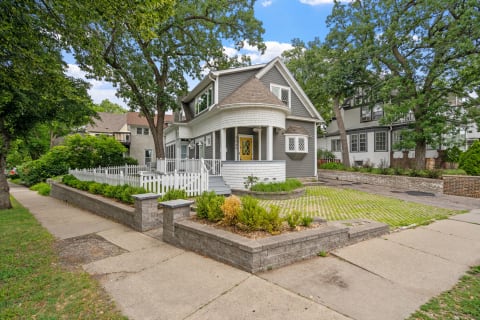 A charming two-story gray house with a white picket fence and a yellow front door in a lush neighborhood.