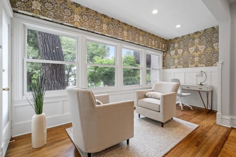 A cozy room with two beige armchairs, large windows revealing greenery, and a desk with a lamp against decorative floral wallpaper.