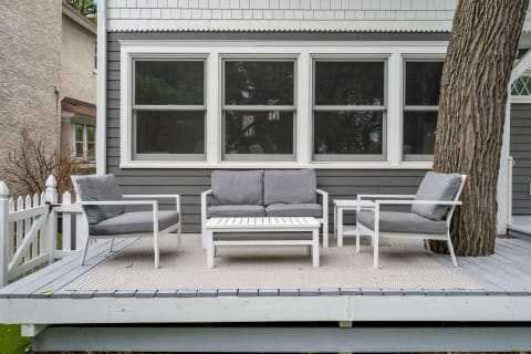 An outdoor patio with gray and white modern furniture, surrounded by greenery and a white picket fence.