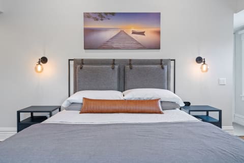 A stylish bedroom featuring a gray upholstered bed, warm lighting, and lakeside sunset artwork.