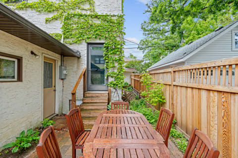 Outdoor wooden dining table with chairs on a brick patio next to a house with ivy on the wall.