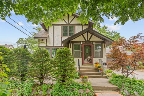 Exterior view of a two-story cream-colored house with dark brown accents, surrounded by greenery and flowers.