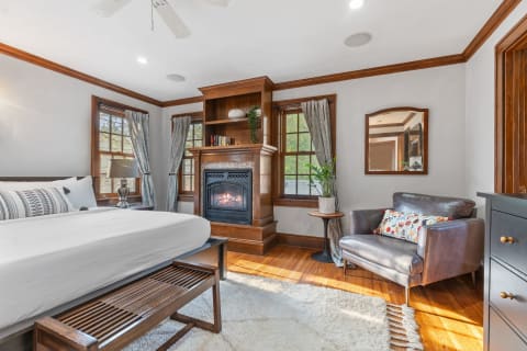 A beautifully styled bedroom featuring a bed, bench, fireplace, and armchair with natural light coming through the windows.