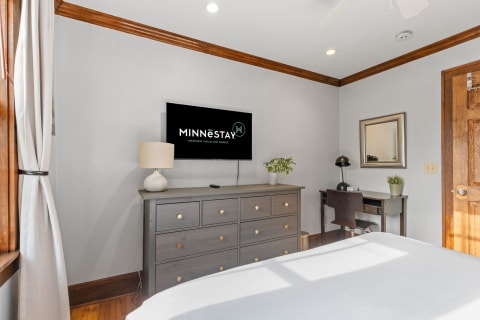 Modern bedroom with a dresser, mounted TV, desk, and natural light.