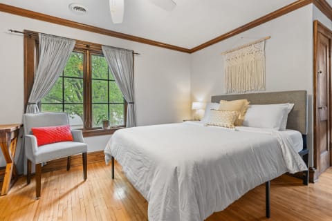 Cozy bedroom featuring a white bed, a gray chair with a red cushion, and a wooden table by the window.