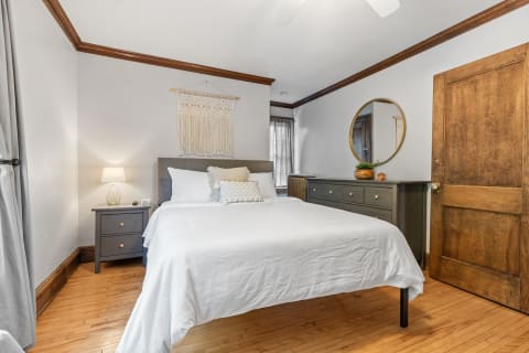A modern bedroom with a white bed, bedside table, a macrame wall hanging, and a dark dresser.