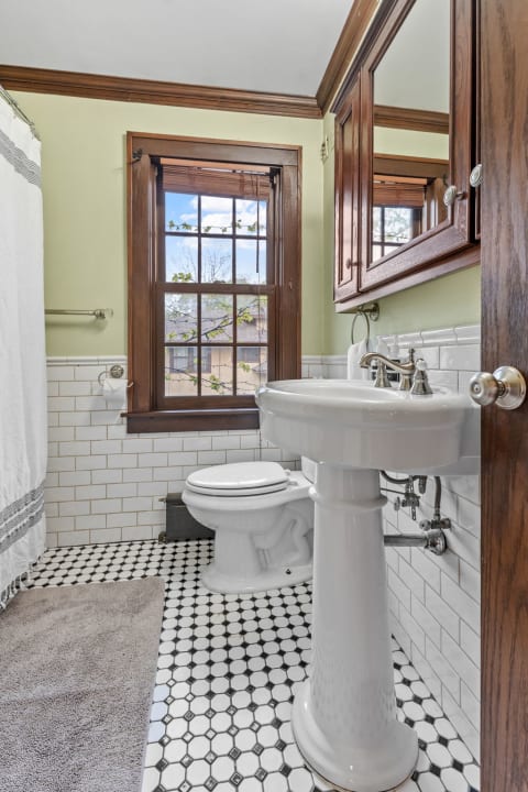 Cozy bathroom featuring a pedestal sink, toilet, and elegant window with wooden trim.