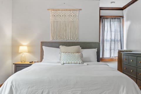 Cozy bedroom with gray headboard, white bedding, macramé wall hanging, and dark dresser.
