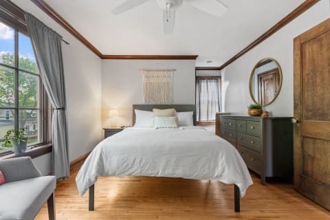 Cozy bedroom with white bedding and a gray upholstered headboard, featuring natural light from a window.