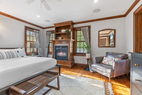A cozy bedroom featuring a bed, armchair, wooden furniture, and a fireplace.