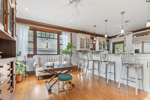 A stylish kitchen and living space featuring a tufted sofa, modern bar stools, natural light, and wooden accents.
