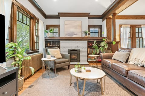 A stylish living room with a leather sofa, armchair, white brick fireplace, and abstract art, filled with natural light and greenery.