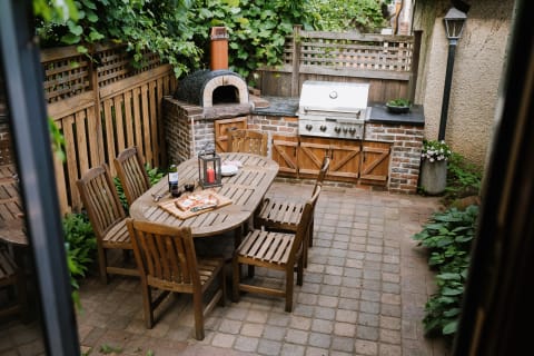 An outdoor dining space featuring a wooden table, chairs, and a barbecue grill surrounded by greenery.